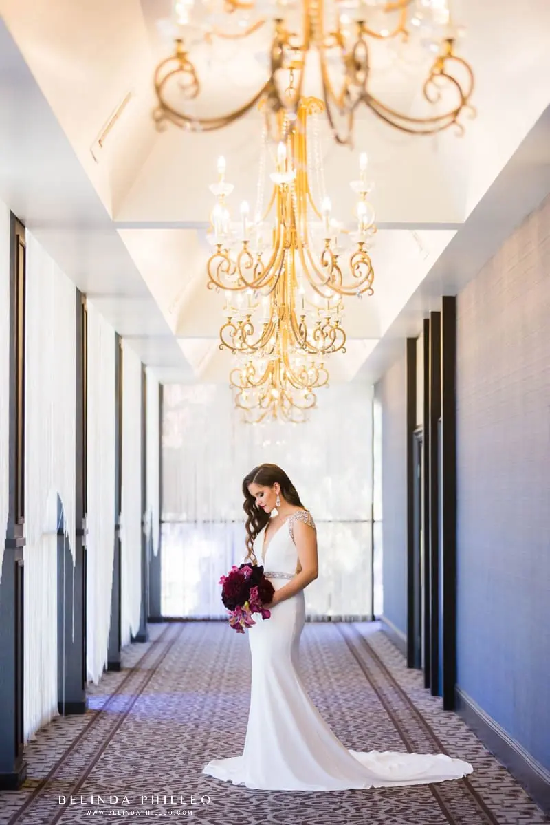 Bride admires her bouquet in a hallway of chandeliers at The Hills Hotel Laguna