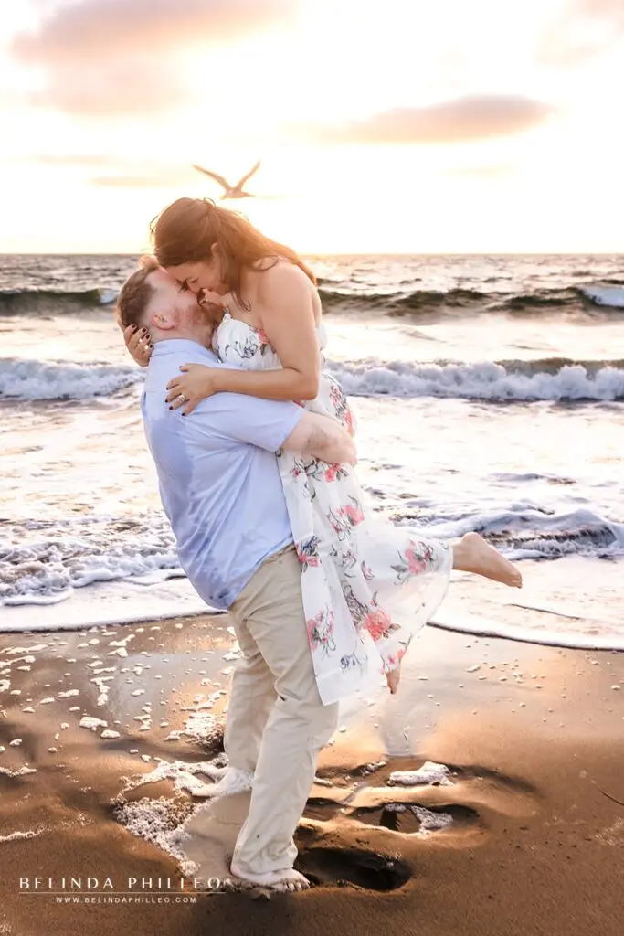 A man picks up his fiance and spins her before giving a kiss on the beach