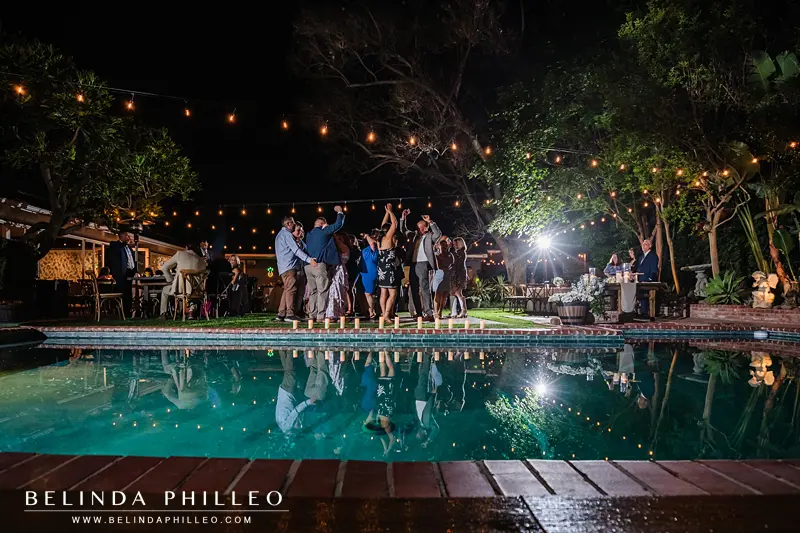 A reflection in a swimming pool shows a crowd dancing the night away at a backyard wedding reception