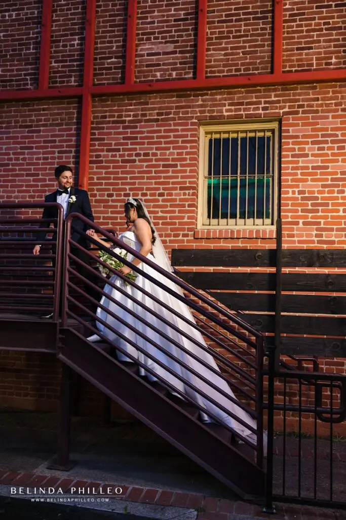 Groom leads his bride by the hand up the stairwell in front of a brick wall