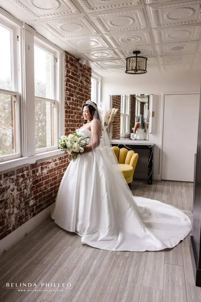 Portrait of a bride standing near a window, admiring her bouquet in the bridal suite at The Charleston in Fullerton, CA
