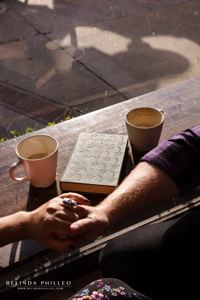 Closeup of engaged couple holding hands next to their coffee mugs and a book, The Betrothed