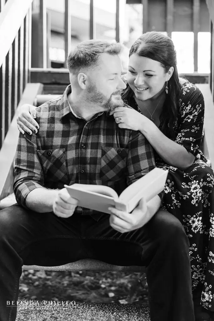 Engaged couple read a book together. The woman leans over the mans shoulder and he looks back lovingly