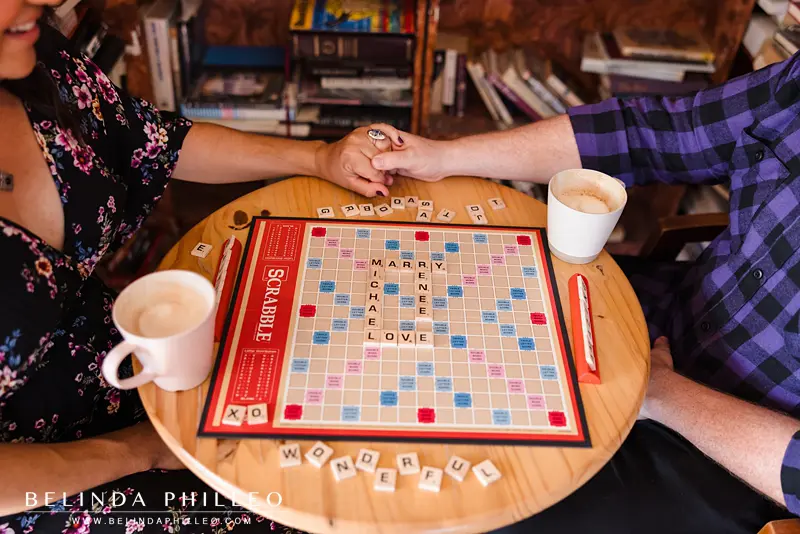Engaged couple hold hands, closeup picture of a scrabble board. The letters spell out "Marry Me" and "love"