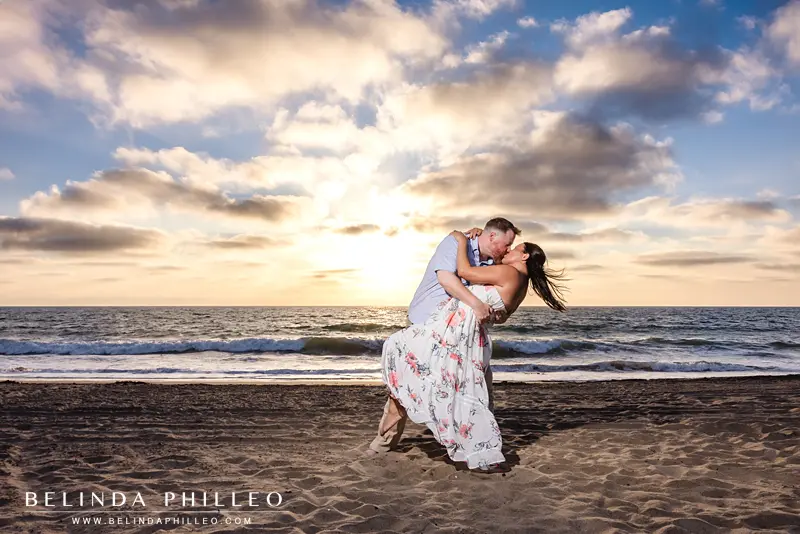 Man dips his fiance and gives her a kiss at sunset on the beach