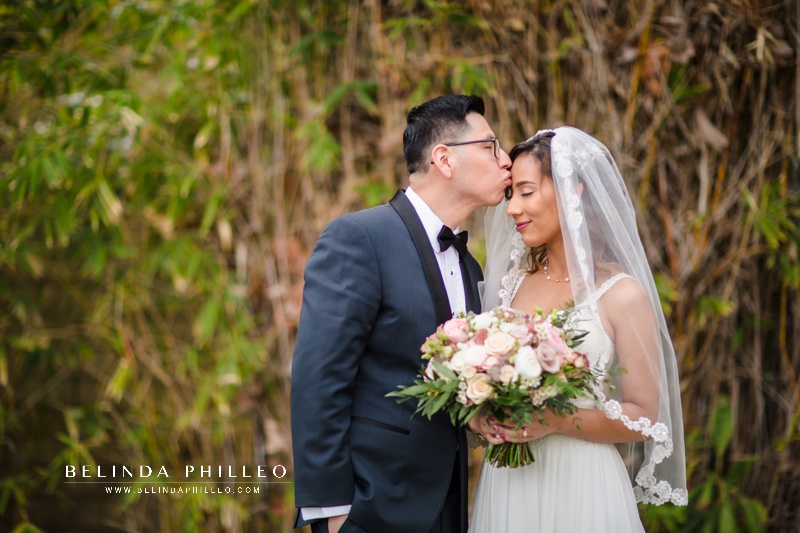 Groom kisses his bride on her forehead while she holds her pastel bridal bouquet