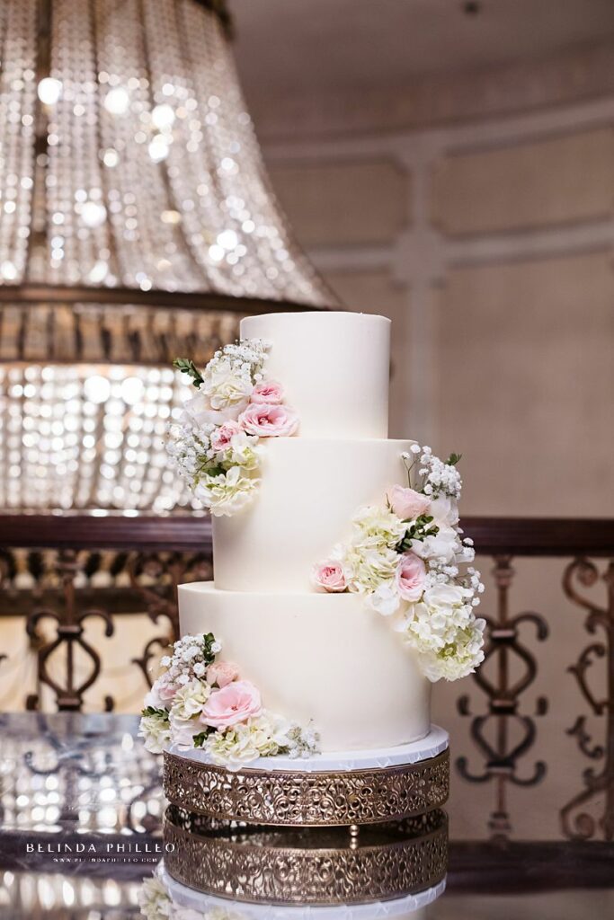 Three tier all white wedding cake with pink rose, white hydrangeas, and baby's breath on an elegant gold cake stand at The Brandview Ballroom in Glendale, CA. Photo by Belinda Philleo